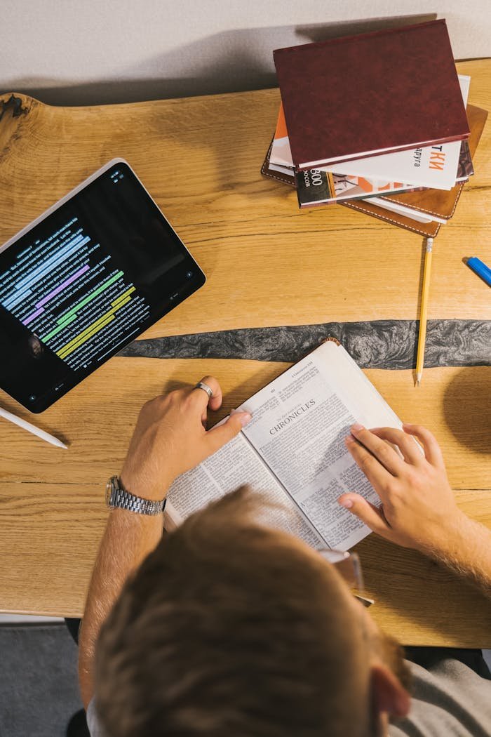 Person reading a Bible at a desk with a tablet and notebooks, emphasizing study and contemplation.